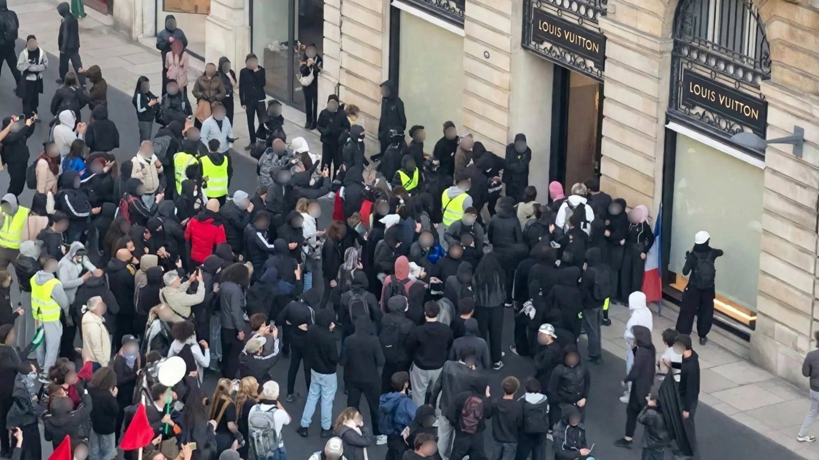 Overhead photo of crowd of protesters, many in black hoods and masks outside luxury store with blurred faces.