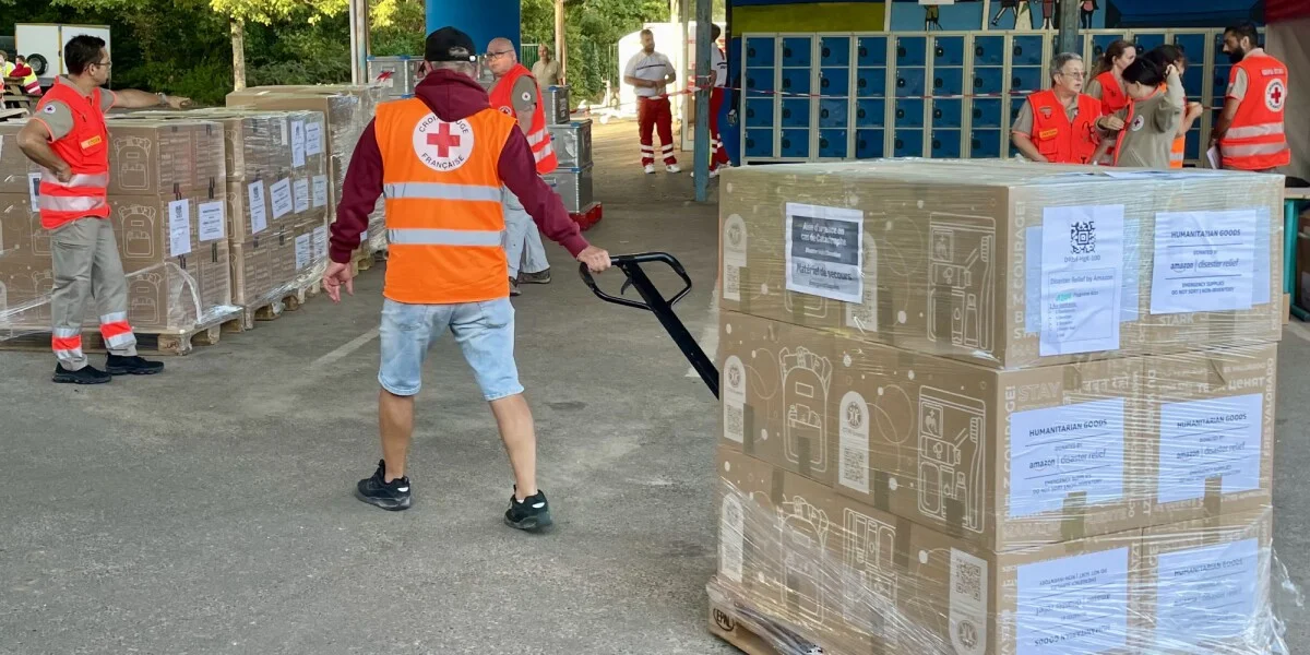 Amazon logo with a backdrop of emergency supplies being prepared for delivery.