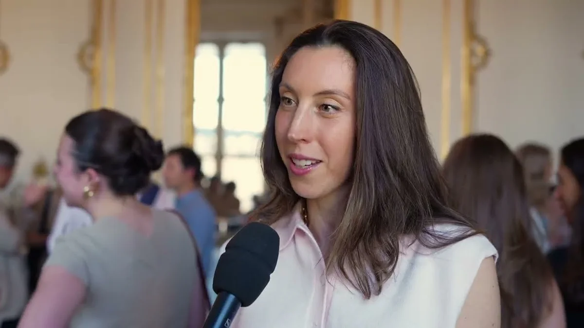 Laura Modiano speaking into a microphone at an event, with other attendees blurred in the background.