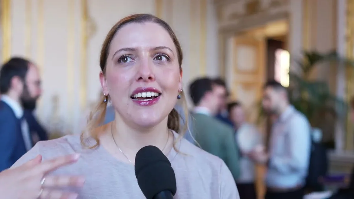 Katia Gil Guzman speaking into a microphone, with blurred attendees in the background.